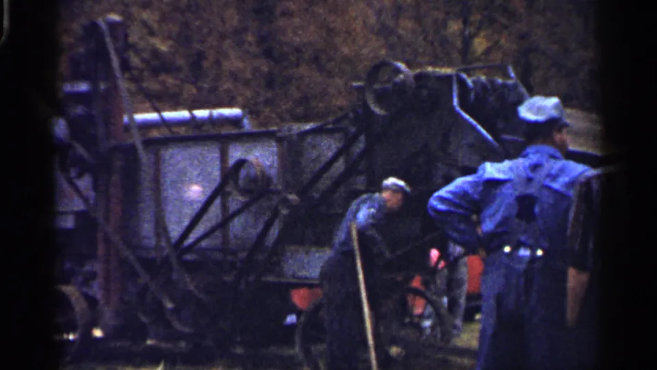 Close 8mm view of men working around pipes, wheels, and metal parts of a steam-powered agricultural machine in Minnesota.