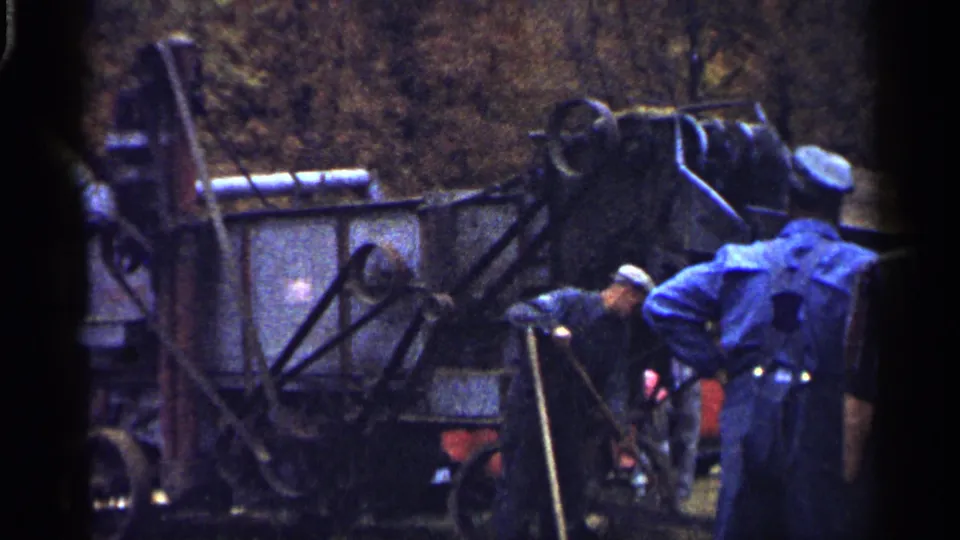 1961 Minnesota frame showing men beside the exposed side of an older agricultural machine with pipes and a large wheel.