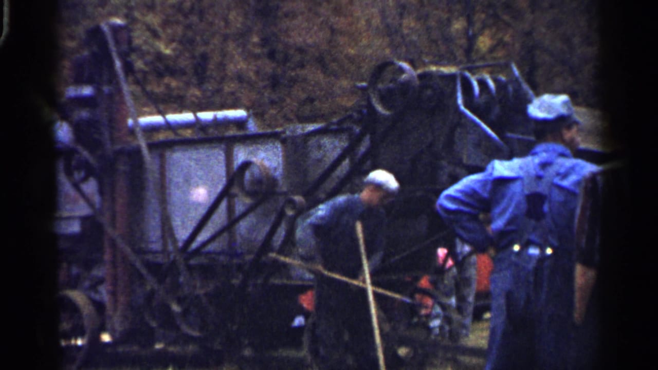Two men in caps work beside a large farm machine in a rural field.