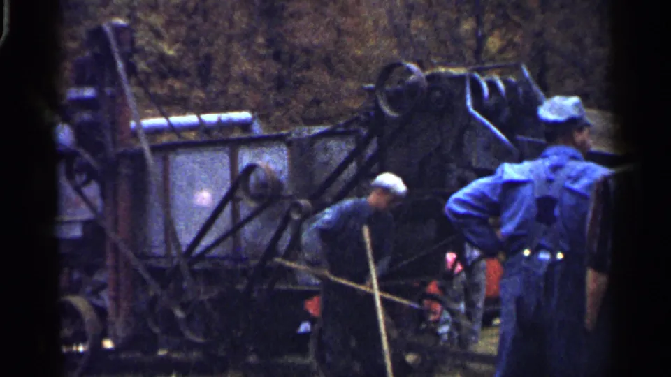 1961 Minnesota home-movie frame showing two adult men in work clothes near a large hay-harvesting machine outdoors.