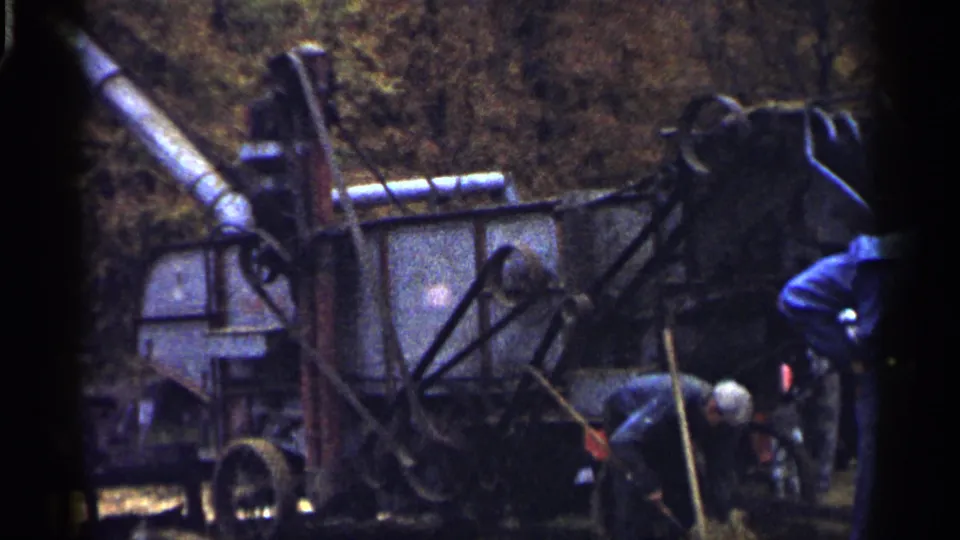 1961 Minnesota home-movie frame showing a worker bent near the base of hay-harvesting equipment, with another figure partly visible.