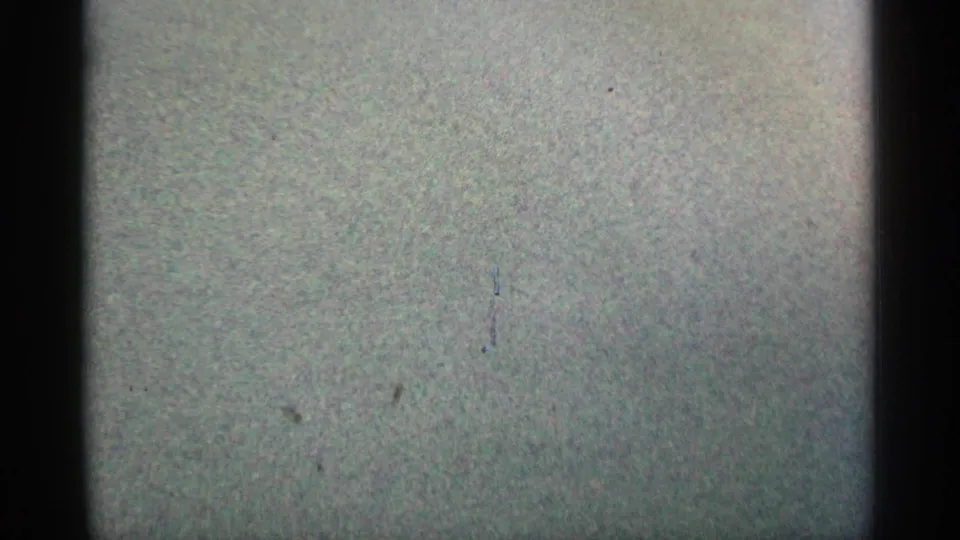 Blank leader-like 8mm frame with heavy grain, pale green-gray exposure, black gate edges, and scattered dust or scratch marks.