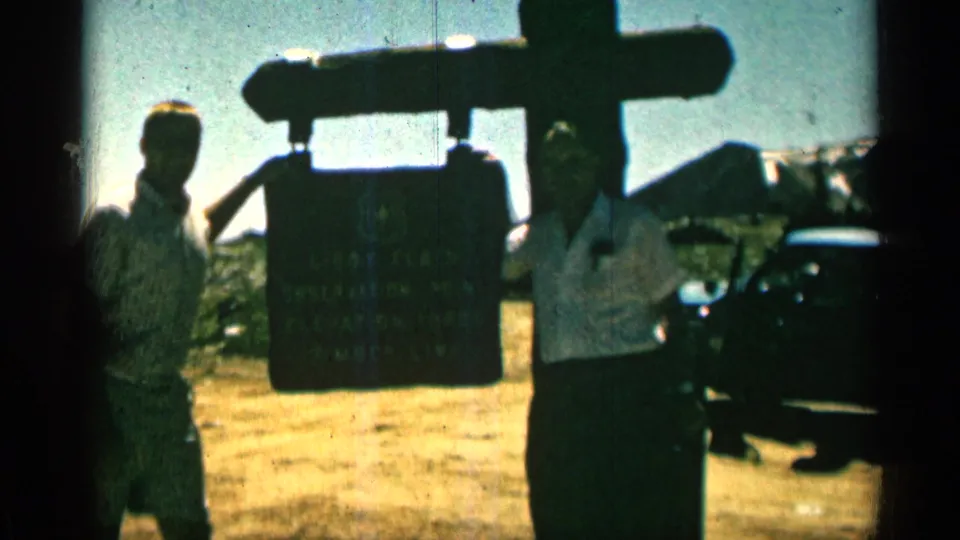 Two adults at a Colorado roadside viewpoint stand near a sign, with mountain scenery and a dark parked car in the background.