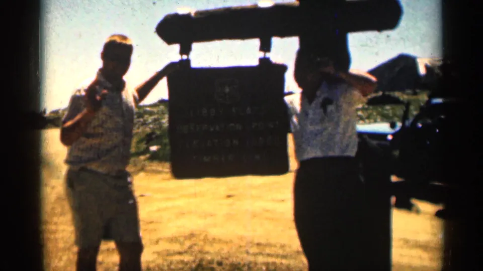 A man waves toward the camera beside the Libby Flats Observation Point sign while another visitor stands near the sign edge.