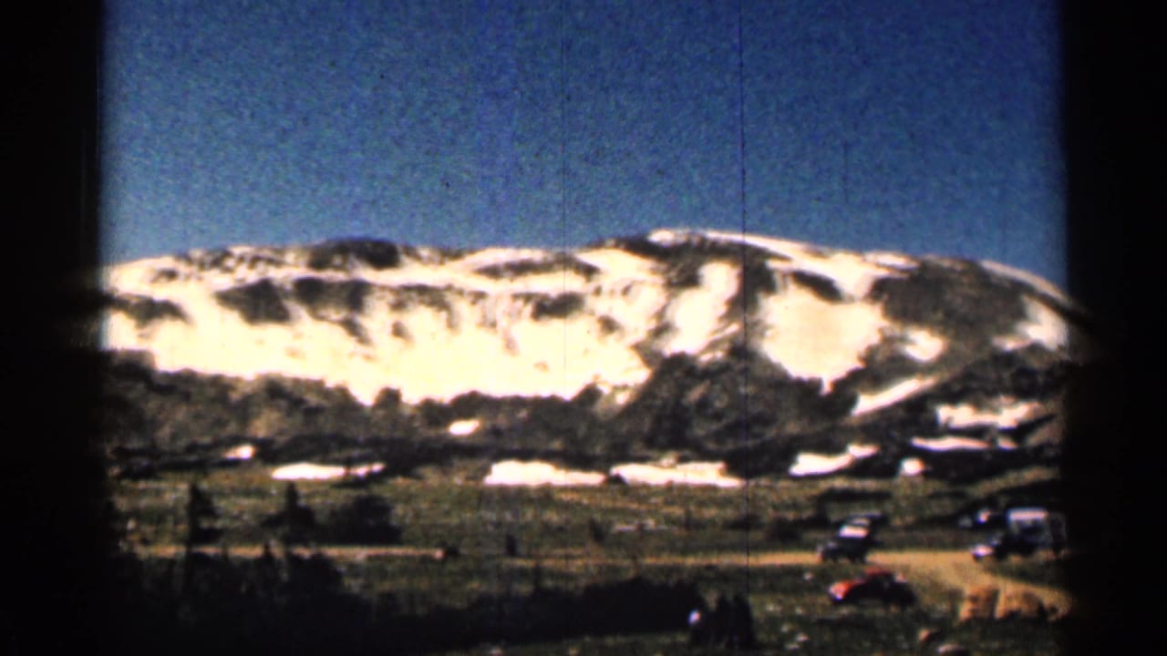 1963 Colorado 8mm frame showing a snow-streaked mountain ridge, open valley ground, distant vehicles, and blue sky.