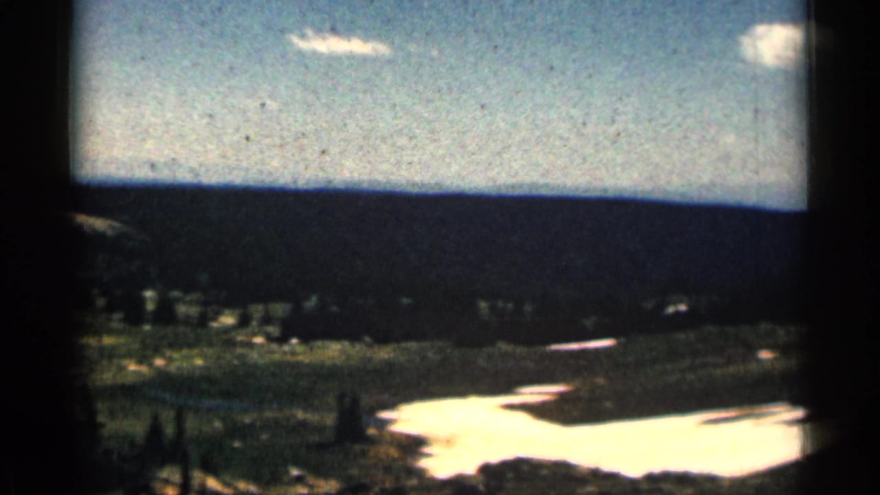 1963 Colorado 8mm frame showing green mountain terrain, lingering snow patches, dark tree lines, and a bright blue sky.
