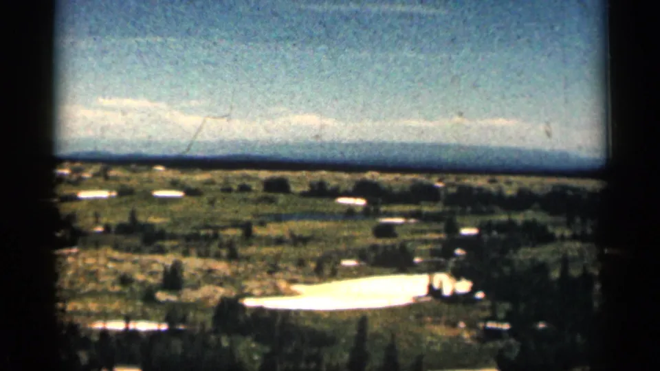 1963 Colorado 8mm frame looking over a green valley-like landscape with snow patches, scattered trees, distant hills, and pale clouds.