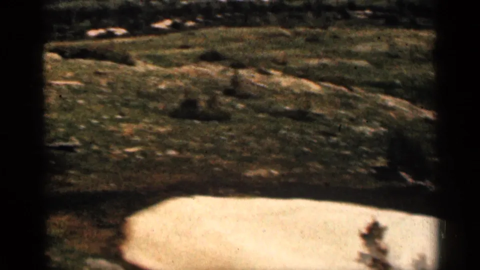 1963 Colorado frame showing a closer rocky grassy slope with a bright foreground snow patch and distant open fields beyond.
