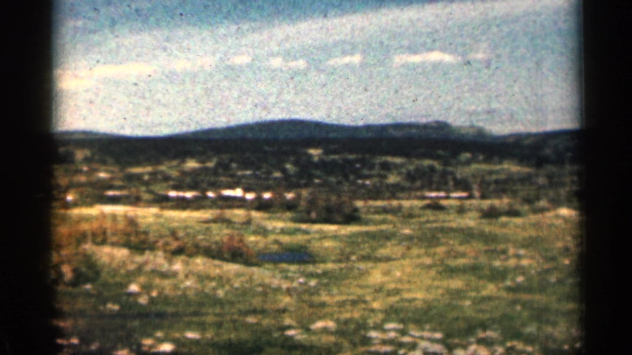 1963 Colorado countryside seen in a grainy 8mm pan, with open grass, rocks, distant hills, and small clouds.