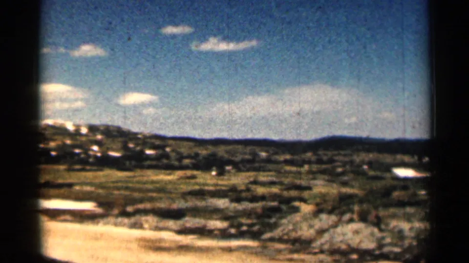 1963 Colorado 8mm frame showing a sunlit dirt patch, rocky grassland, low hills, and bright sky with small clouds.