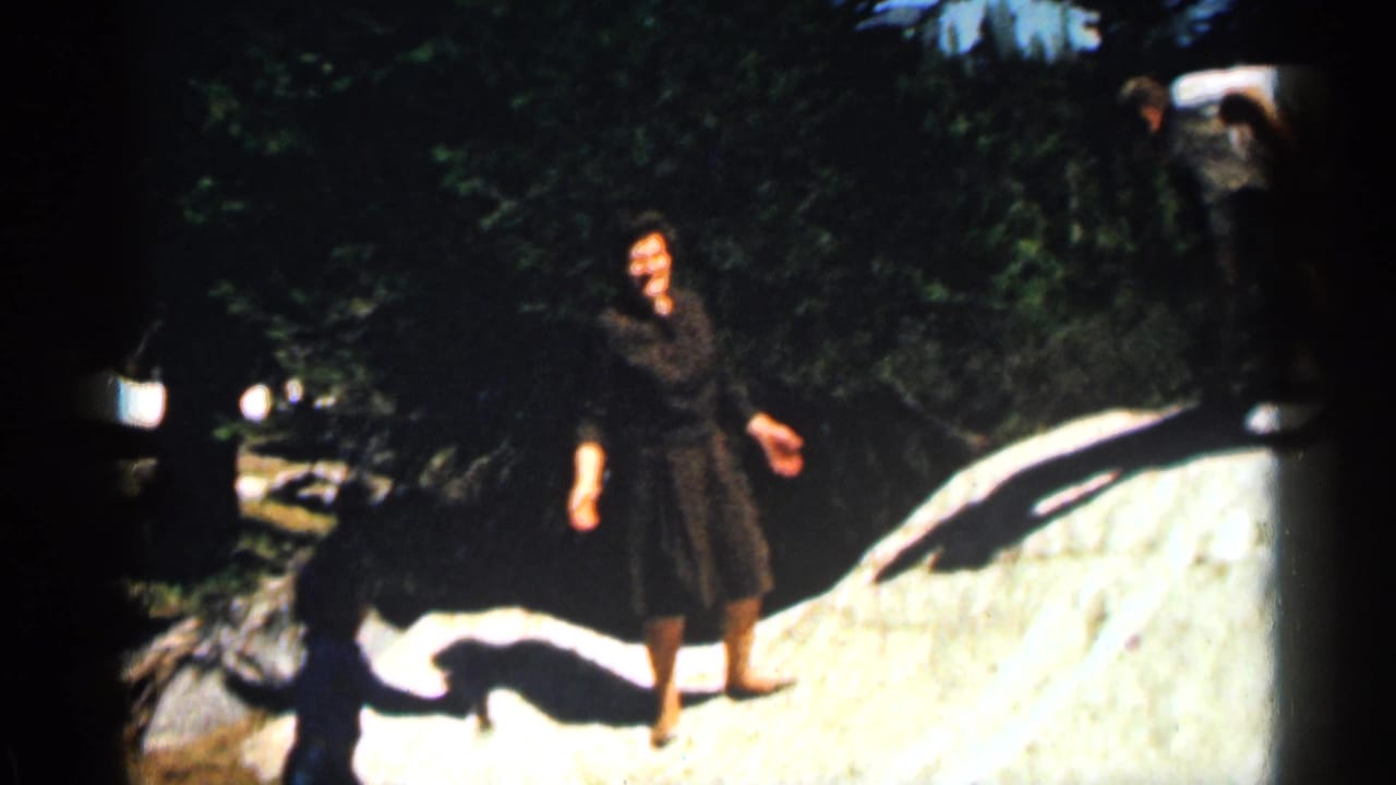 Woman in a dark dress stands near a pale rock in a forested outdoor area.