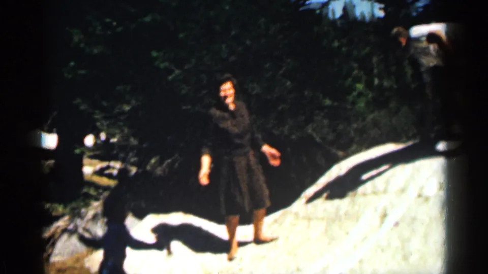 1963 Colorado home-movie frame showing a woman in a dark dress near a pale rock and trees.