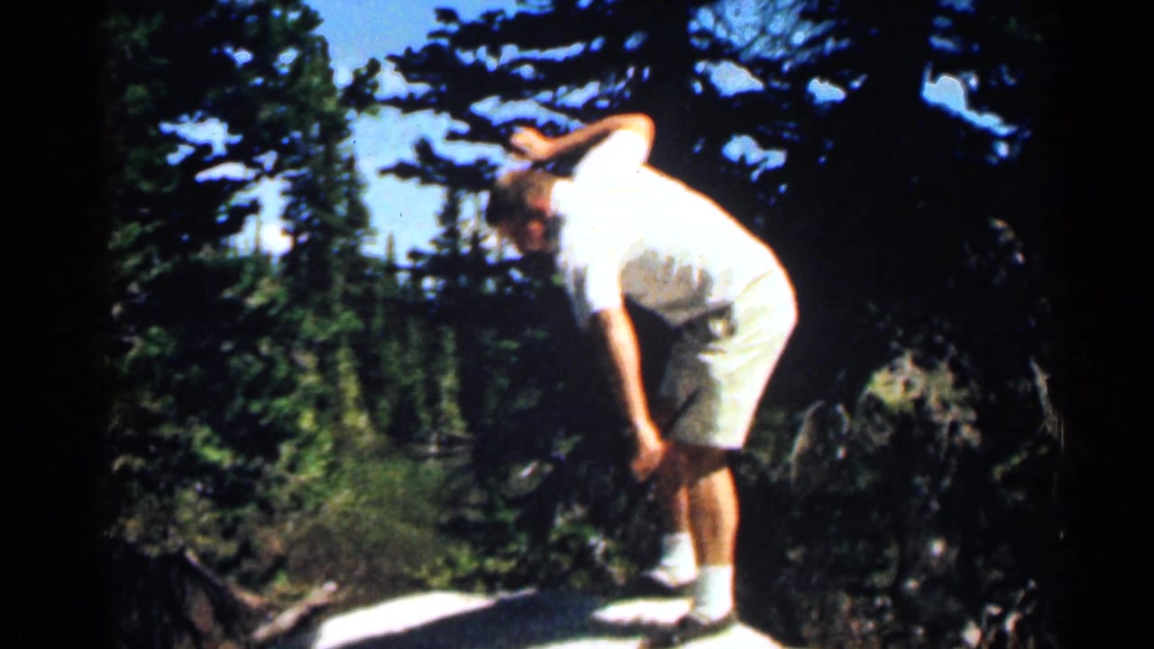 Man in a white shirt bends forward on a sloped rock among trees.