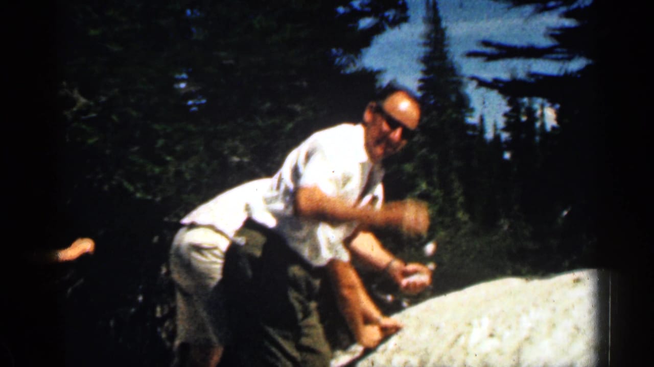 1963 Colorado home-movie frame of a man leaning over a snow patch while family members stand nearby on a mountain slope.