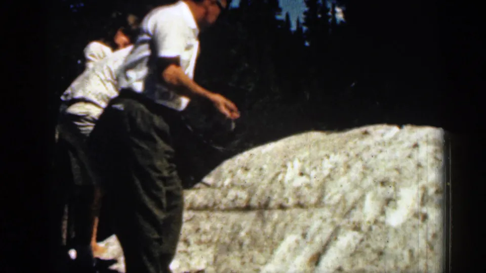 1963 Colorado home-movie still of a man turning near the snow as another adult stands close behind on the grassy slope.