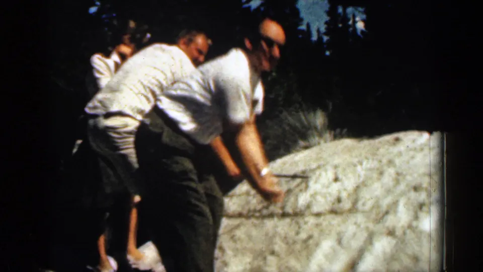 1963 Colorado frame of the man bending at the snow patch, with relatives clustered nearby against evergreen trees.