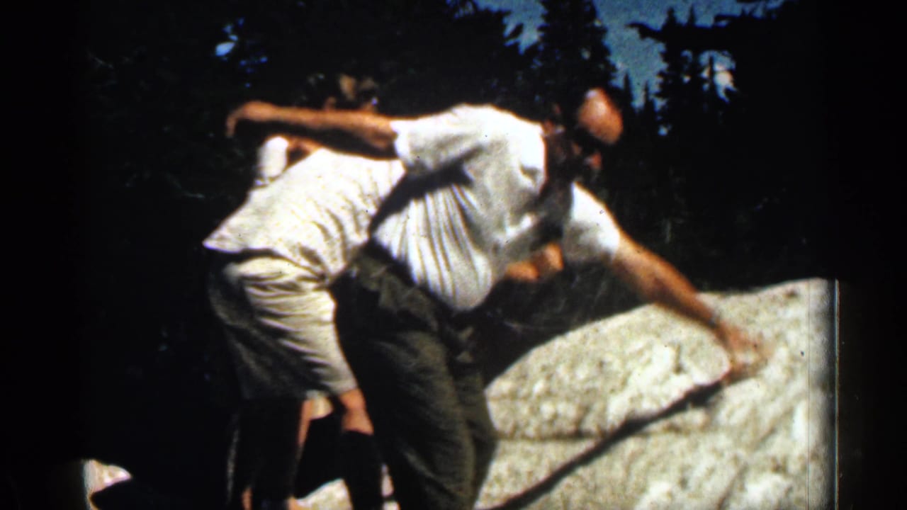 1963 Colorado home-movie frame of two men in light summer clothing moving over a large rock in a forest.