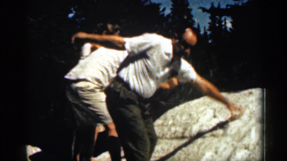 1963 Colorado home-movie frame of two men in light summer clothing moving over a large rock in a forest.