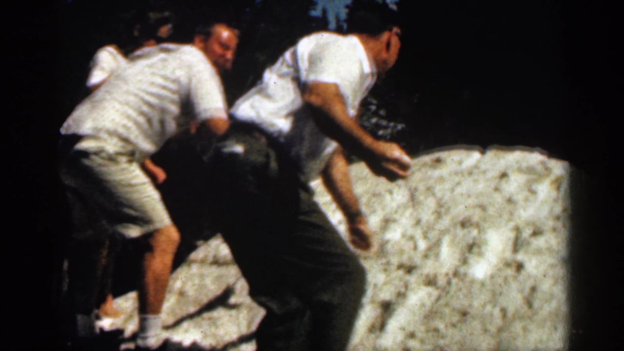 Two men in casual 1960s-style summer clothing climb across a pale rock in a wooded Colorado setting.
