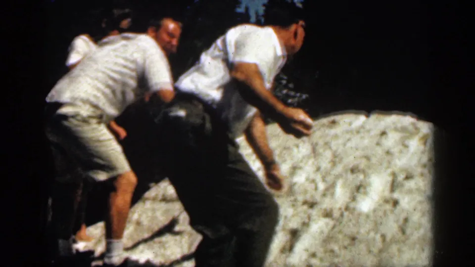Two men in casual 1960s-style summer clothing climb across a pale rock in a wooded Colorado setting.