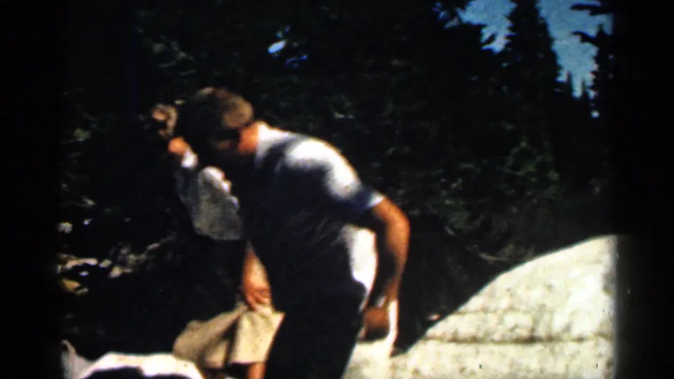 A man passes in front of a woman and pale rocks in a wooded Colorado hiking scene from 1963 home-movie footage.