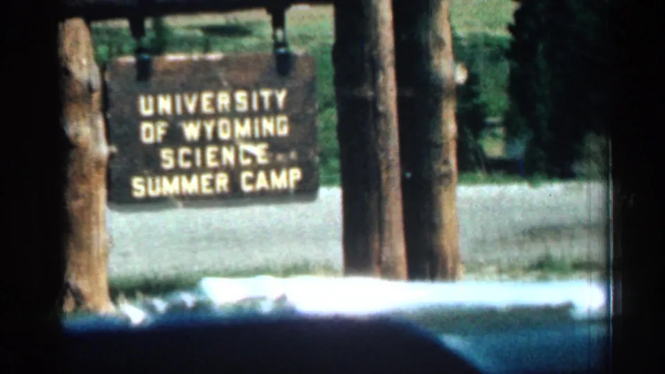 8mm frame of a wooden University of Wyoming Science Summer Camp sign standing by trees and a road or path in an outdoor camp setting.