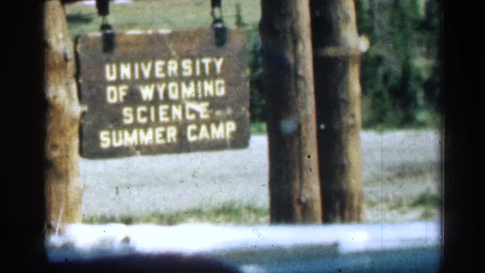 1963 Colorado 8mm frame showing a wooden University of Wyoming Science Summer Camp sign near a roadside or camp entrance.