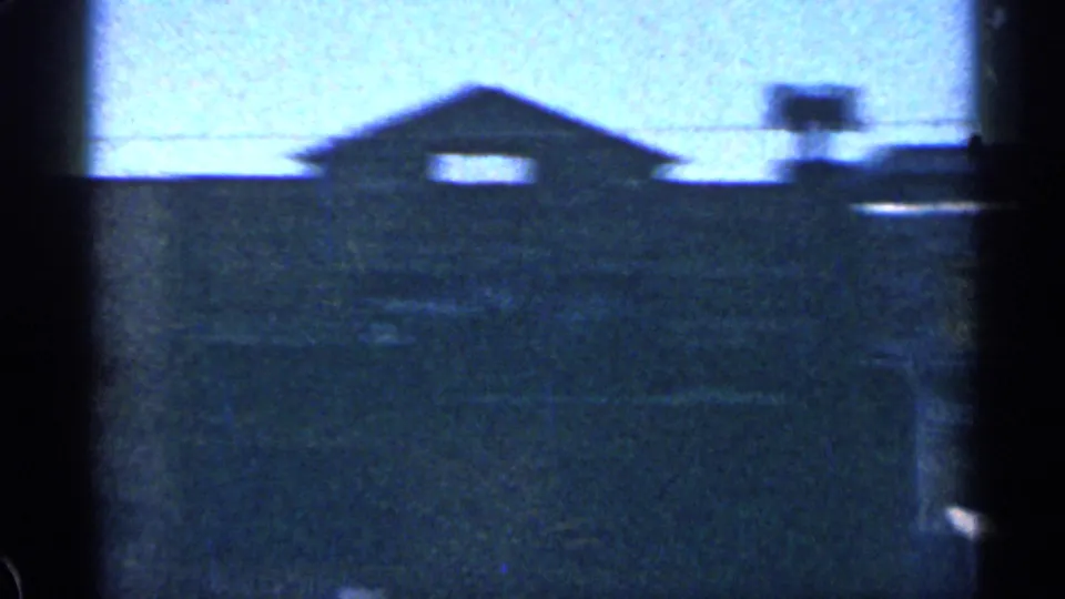 Low-light 1963 Colorado home-movie frame with a dark gabled building, small lit windows, and overhead lines.