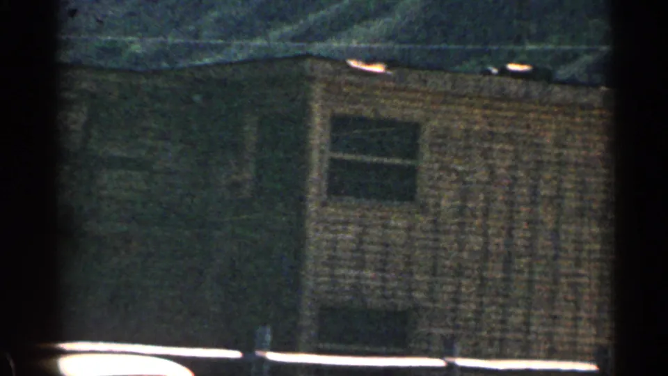 1963 Colorado 8mm frame showing the side of a rustic building with small windows, shingled wall texture, and a moving-vehicle viewpoint.