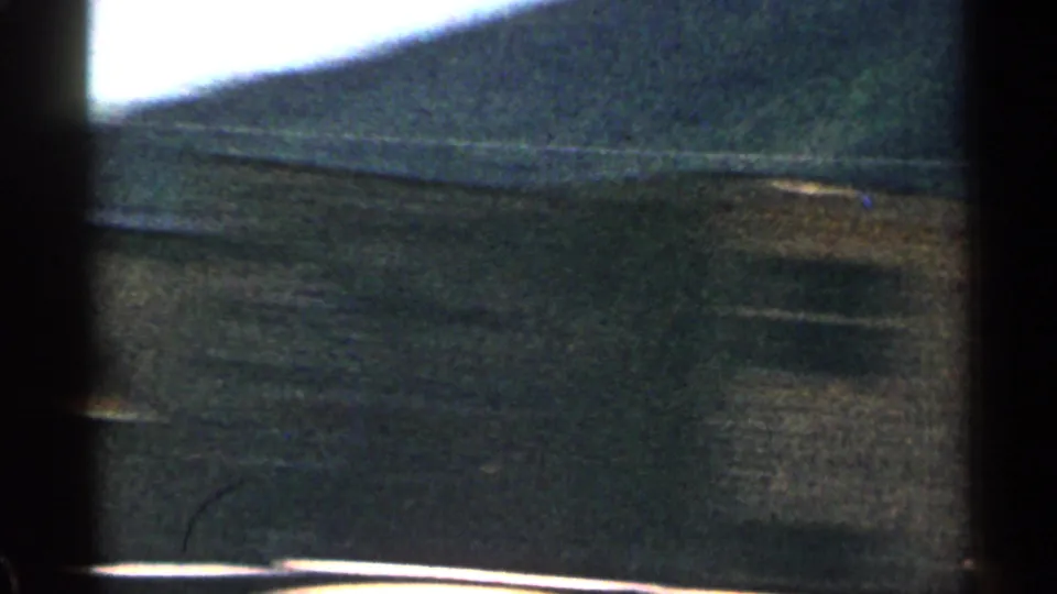 1963 Colorado 8mm frame with a dark station building edge, mountain slope, bright sky, and a blurred vehicle roof crossing the lower frame.
