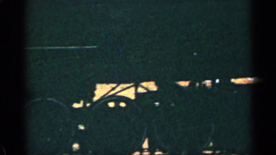 1963 home-movie frame showing the lower locomotive body and wheels as a dark silhouette over sunlit trackside ground.