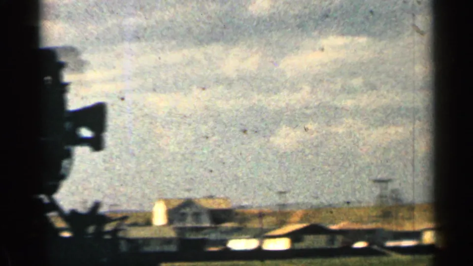 1963 Colorado 8mm frame showing a cloudy sky, low rural structures, and a dark foreground silhouette at the left edge.