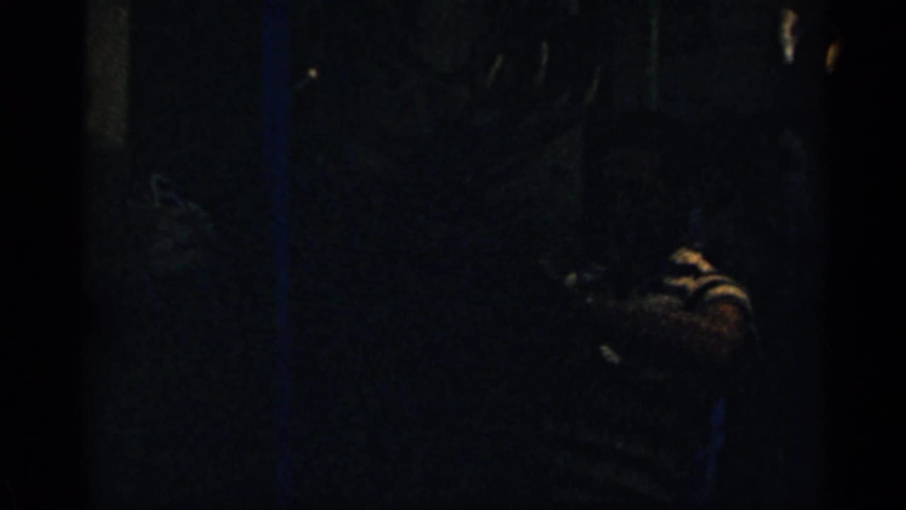 Very dark interior frame with a faint figure in a striped shirt near the right side.