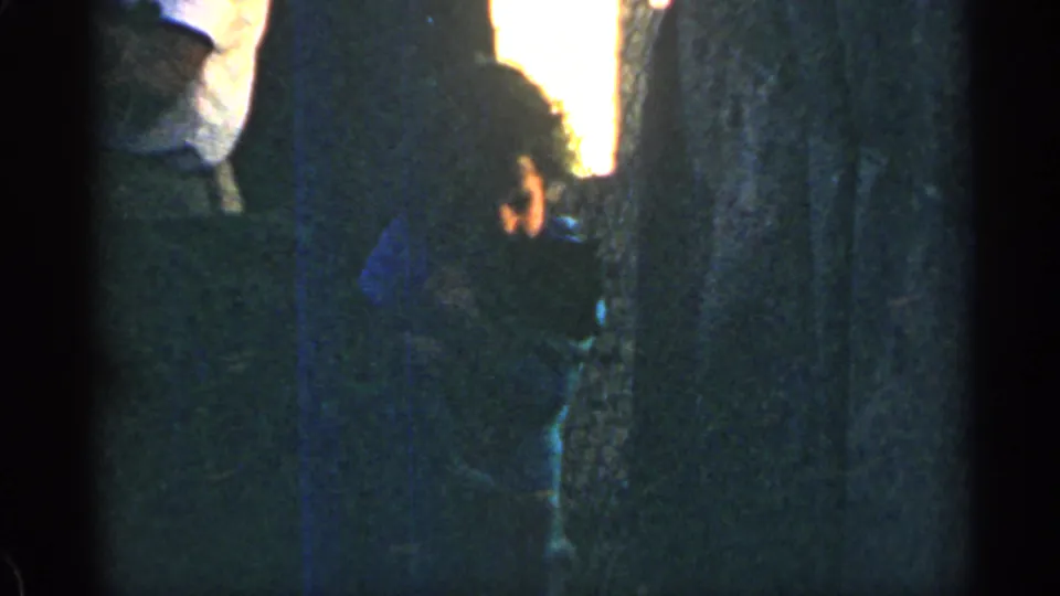 1963 Colorado 8mm frame of a small child in a blue top holding a puppy near tree trunks in dim outdoor light.