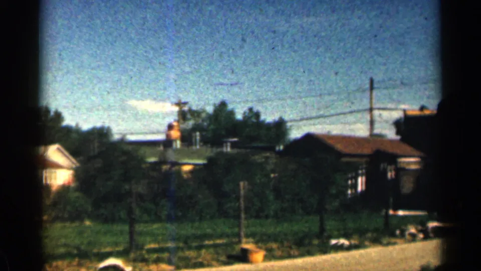 1963 Colorado 8mm frame widening to show the tower near rooftops, trees, utility poles, grass, and roadside pavement.