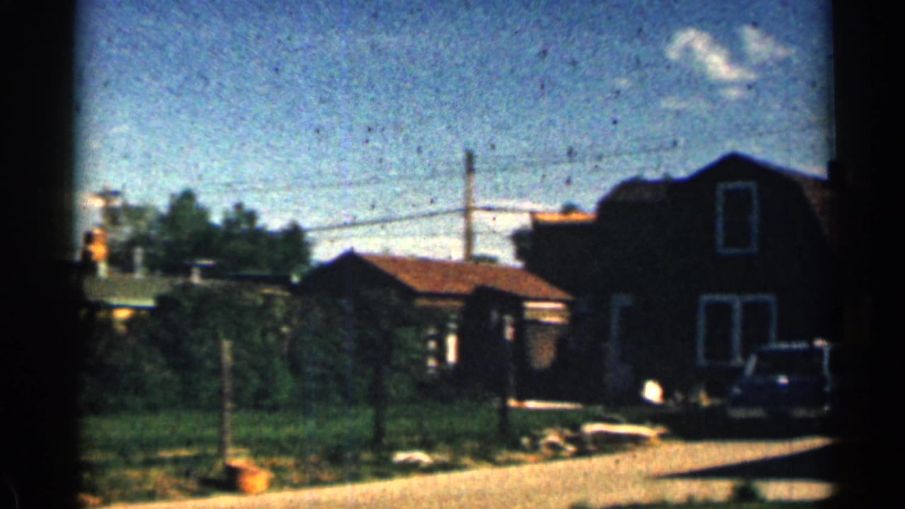 1963 Colorado home-movie frame showing a sunlit residential street with modest houses, lawns, overhead wires, and a parked car.
