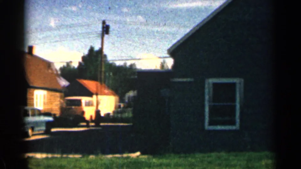 Colorado home-movie frame showing house exteriors along a street, a pale parked vehicle, utility lines, and a dark foreground wall.