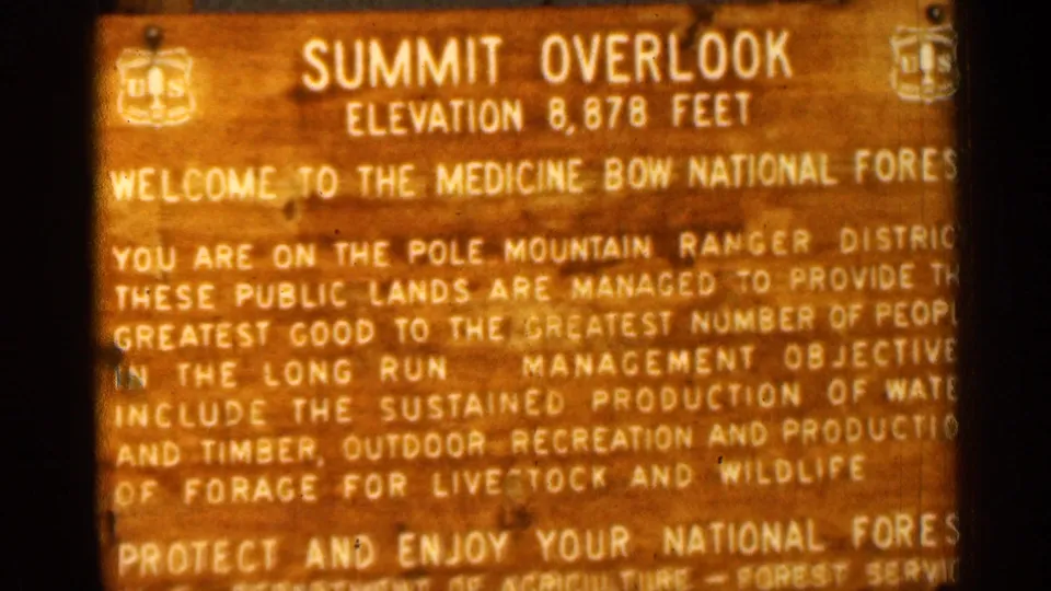 1963 home-movie frame of the Summit Overlook sign, with Pole Mountain Ranger District text visible on the weathered wooden board.