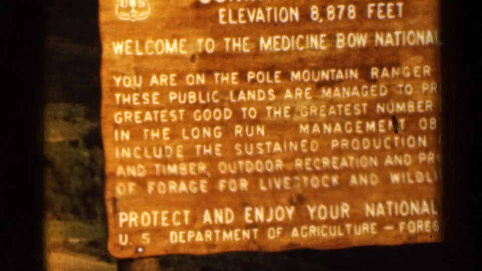 1963 home-movie frame showing the lower half of the Summit Overlook sign, including recreation, water, timber, forage, and Forest Service wording.