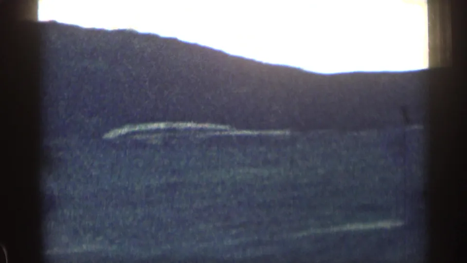 Vehicle-window view of a dark Colorado hillside and open terrain, with bright sky above and soft 8mm film grain across the frame.