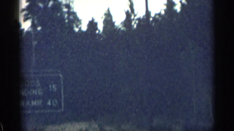 1963 Colorado 8mm frame showing a partly blurred roadside distance sign near evergreen trees during a cloudy mountain drive.