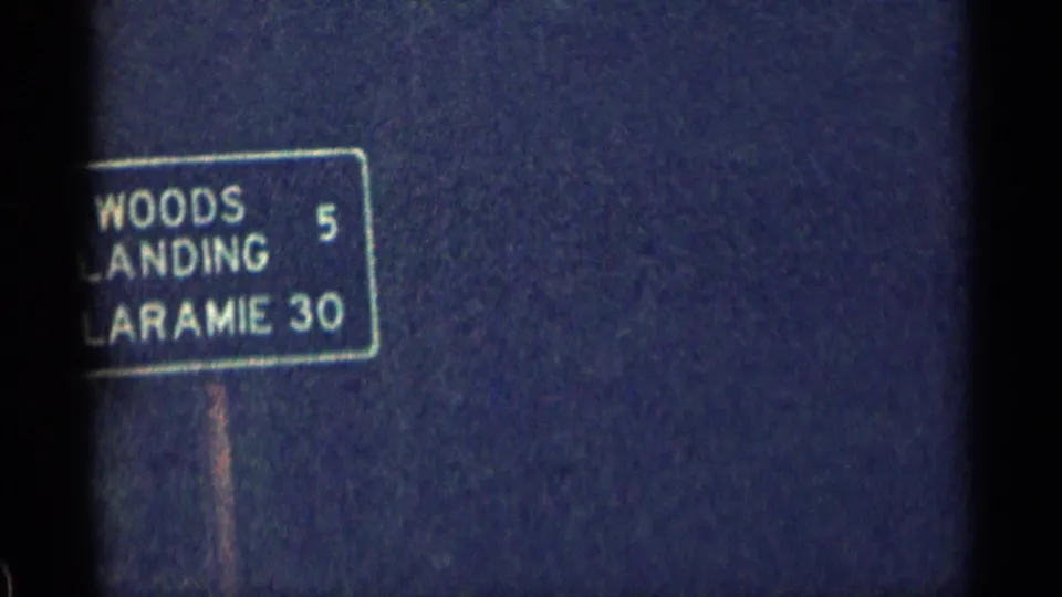 1963 Colorado 8mm frame with a readable sign for Woods Landing and Laramie, framed against trees and cloudy light.