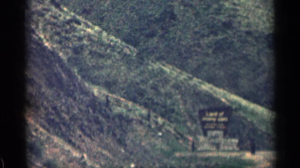 1963 Colorado 8mm frame showing a sloped wooded roadside and a small lodging sign below the hillside.