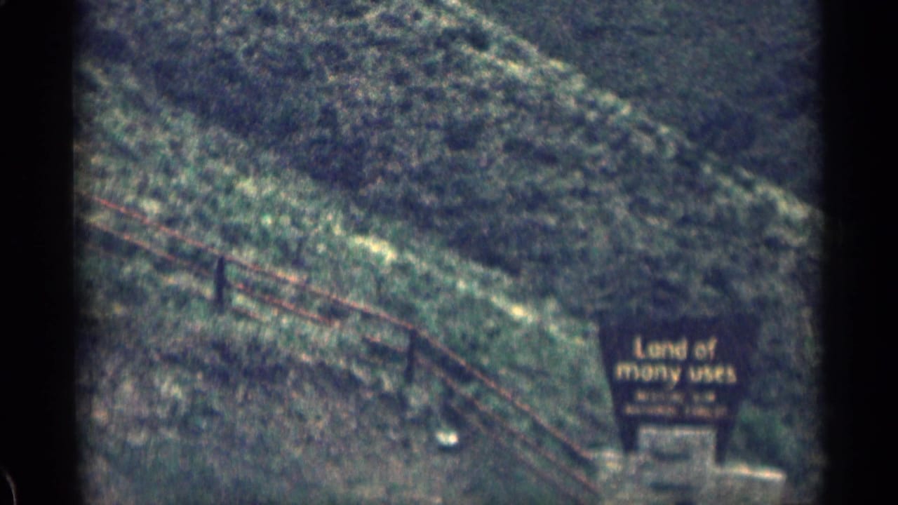 1963 Colorado 8mm frame showing a grassy mountain slope, roadside fence, and a partially legible sign reading Land of many uses beside the road.