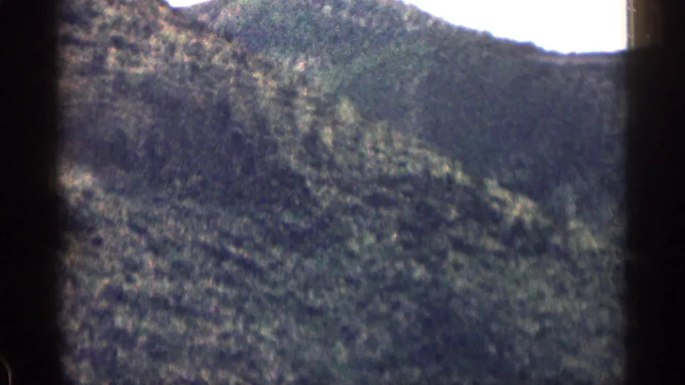 1963 Colorado home-movie frame with a brushy mountain slope, pale sky, and visible film gate shadow.