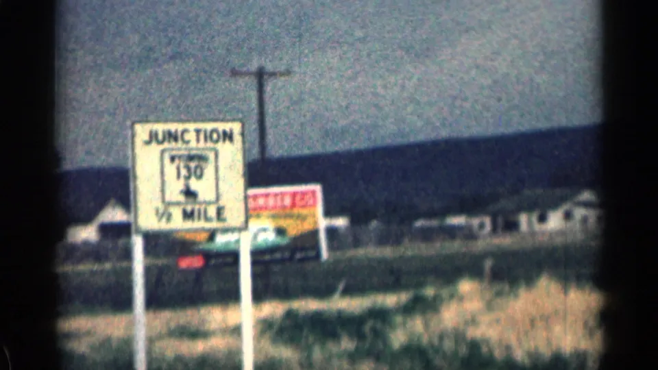 1963 Colorado roadside frame with a junction sign in front of sparse buildings, utility poles, and low hills.
