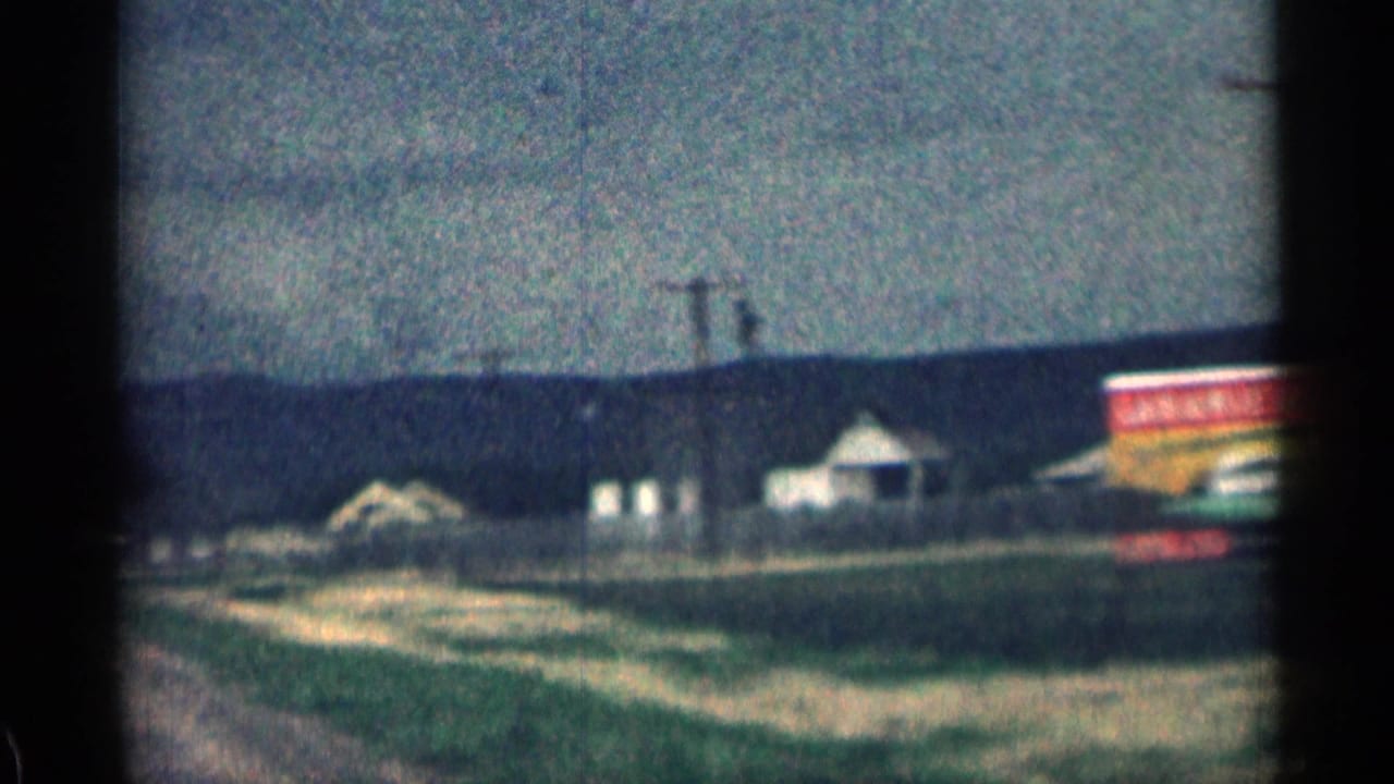 Rural roadside with low buildings, a large sign, utility poles, and hills beyond.