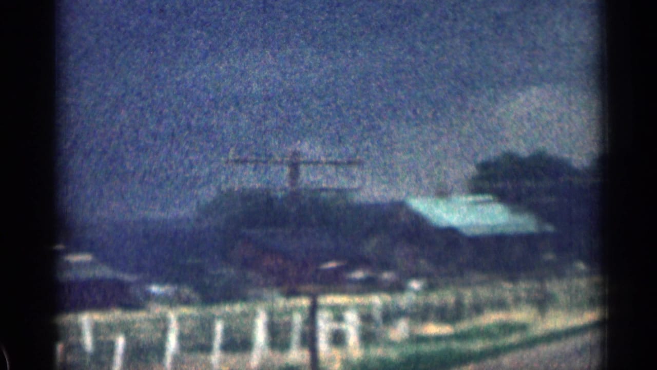Roadside buildings and fencing with a pale roof, trees, and hills in the background.