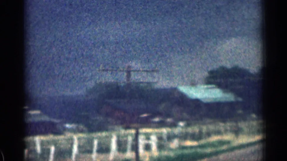 Soft 1963 Colorado roadside view with fenced ground, small buildings, a pale roof, trees, and distant hills.