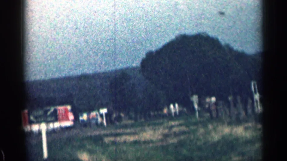 Grainy 1963 Colorado frame with roadside posts, trees, a red sign or structure, rough ground, and hills beyond.